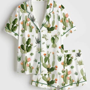 May include: White pajama set featuring a cactus print. The short-sleeved top and shorts have a green trim. The cactus design includes green cacti with orange flowers and various succulent plants.