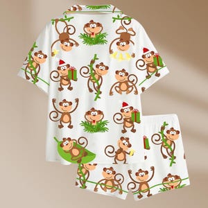 May include: White pajama set with a playful monkey print. The short-sleeved top and shorts feature cartoon monkeys in various poses, some holding gifts or bananas, set against a backdrop of green vines and grass.