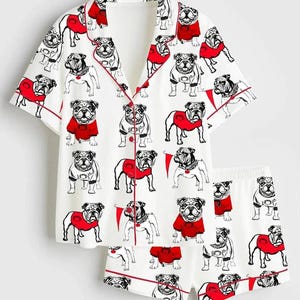 May include: White pajama set with a repeating pattern of bulldog illustrations wearing red sweaters. The short-sleeved top and shorts are trimmed with red piping.