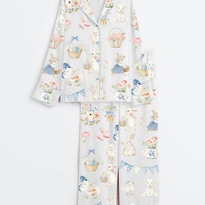 Op de afbeelding: Een lichtgrijze pyjamaset met een grillige paasprint. De set bestaat uit een overhemd met knopen en een broek, versierd met konijnen, lammeren, vlinders, bloemen en manden. De randen zijn afgewerkt met een blauw ruitpatroon.