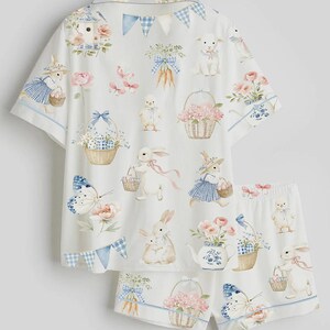 Op de afbeelding: Witte pyjamaset met een grillige paasprint. De set bevat een top met korte mouwen en een short, beide versierd met konijntjes, bloemenmanden, vlinders en wortels. Blauwe en roze accenten en afwerkingen maken het ontwerp compleet.