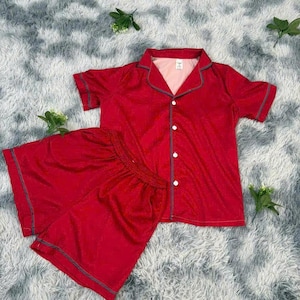 May include: A red pajama set featuring a short-sleeved button-down shirt and matching shorts. The shirt has a collar and gray trim, with white buttons down the front. The shorts have a drawstring waist and gray trim. The set is made of a soft, silky material.