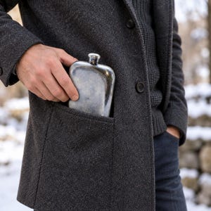 May include: A silver-toned metal flask with a smooth, rounded rectangular shape and a small screw-top cap. The flask is partially inserted into the deep pocket of a dark gray wool-blend coat, showing its metallic finish and curved edges.