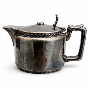 May include: A silver-colored teapot with a lid and handle. The teapot has a spout for pouring and a decorative rope-like design around the base and lid. The handle is curved and the lid has a small knob.