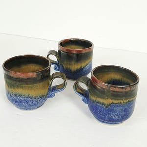 May include: Three ceramic mugs featuring a reactive glaze with a color gradient transitioning from a deep blue base to a mustard yellow middle and a dark brown rim. Each mug has a curved handle and a smooth, rounded body shape with a glossy finish.