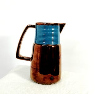 May include: A pitcher featuring a metallic copper-toned lower body and a contrasting ribbed upper section in a vibrant blue glaze. The vessel includes a single angular handle on the side and a small pouring spout at the top rim of the container.