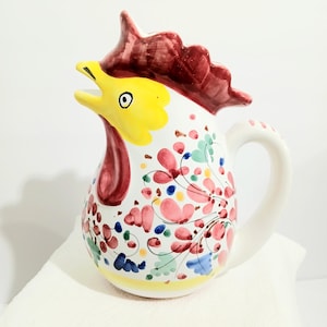 May include: A white ceramic pitcher in the shape of a rooster, featuring a yellow head, red comb and wattle, and a floral pattern in pink, red, blue, and green. The pitcher has a curved handle and a yellow base.