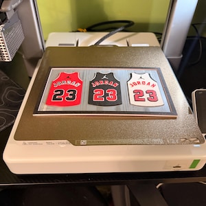 May include: A wooden sign featuring three basketball jerseys in red, black, and white, each with the number 23 and the name "Jordan." The jerseys are displayed on a light-colored surface.