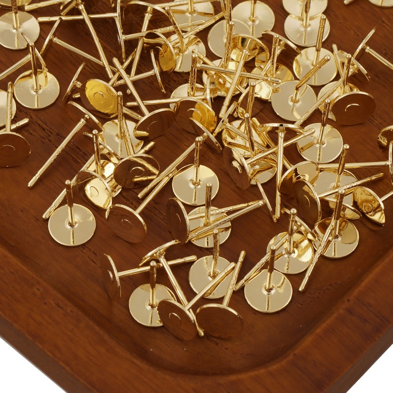 May include: A close-up of a pile of gold-toned earring posts and backs. The earring posts are round and have a flat back. The earring backs are small and round.