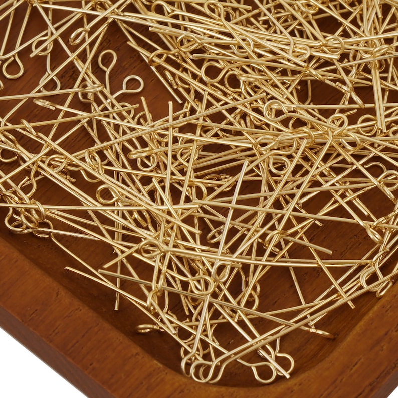 May include: A large quantity of gold-colored head pins with loops on one end. The pins are scattered in a wooden tray.