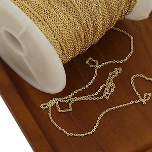 May include: A spool of gold chain, with a length of chain laid out on a wooden surface. The chain is a fine, delicate style.