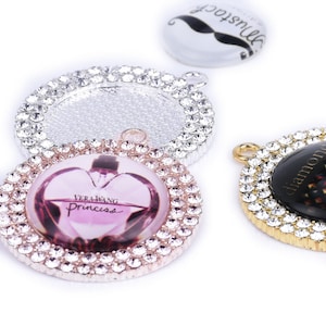 May include: Three round, silver-toned, rhinestone-encrusted pendants. One pendant features a clear glass insert with a black mustache design and the word "mustache" written in cursive. Another pendant features a clear glass insert with a pink and white image of a perfume bottle with the text "Vera Wang Princess". The third pendant features a black glass insert with a gold design and the word "diamond" written in gold.
