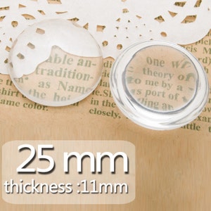 Può includere: Due cabochon in vetro trasparente, uno di 25 mm di diametro e 11 mm di spessore, l'altro leggermente più piccolo. I cabochon sono appoggiati su una superficie marrone con testo visibile sullo sfondo.