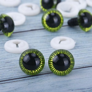 May include: A close-up of two green plastic safety eyes with black pupils. The eyes have a gold glitter finish and are surrounded by a ring of green.