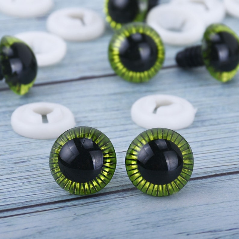 May include: A close-up of two green plastic safety eyes with black pupils. The eyes have a gold glitter finish and are surrounded by a ring of green.