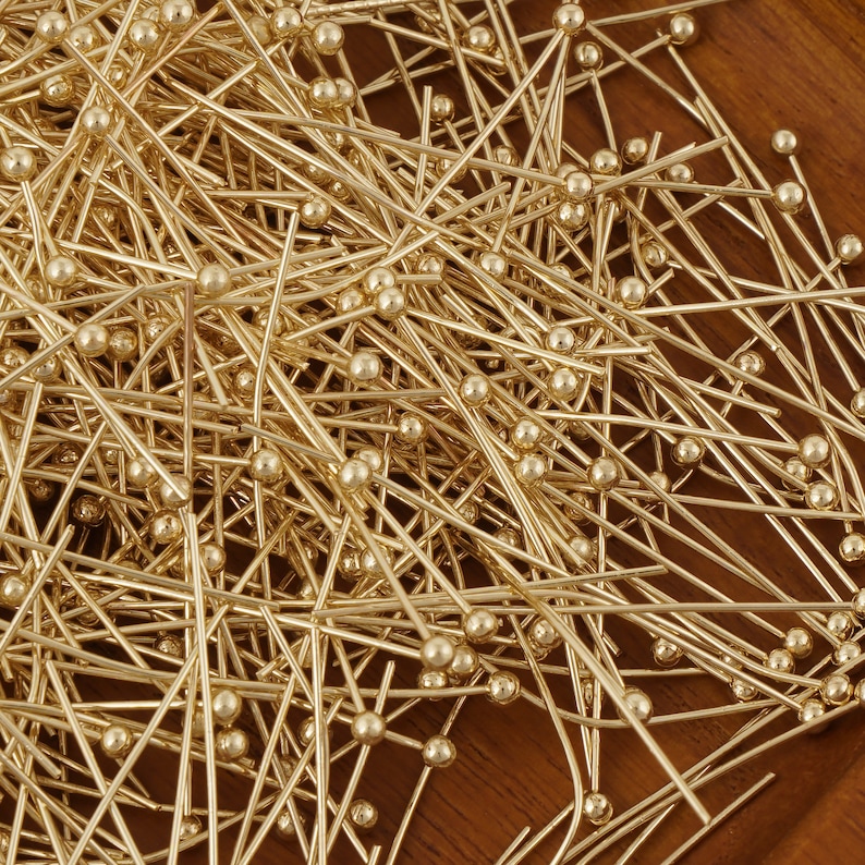 K&ouml;nnte beinhalten: Ein gro&szlig;er Haufen goldfarbener gerader Stecknadeln mit runden K&ouml;pfen. Die Nadeln liegen auf einer Holzoberfl&auml;che verstreut.