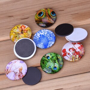May include: Assortment of round glass magnets with various designs, including peacocks, fairies, and floral patterns. The magnets have a black backing.