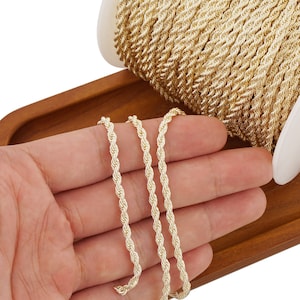 Könnte beinhalten: Goldene Kordelkette, eine Art von Dekorband, das für Bastelarbeiten und Schmuckherstellung verwendet wird. Die Kette ist auf einer Holzoberfläche und in einer Hand zu sehen.