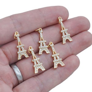 Op de afbeelding: Vijf goudkleurige Eiffel toren bedels bezet met heldere strass steentjes. De bedels zijn gedetailleerd met de structuur van de iconische toren en hebben een lus aan de bovenkant om aan sieraden of andere knutselwerken te bevestigen.
