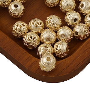 May include: A close-up of a wooden tray filled with gold-coloured metal beads. The beads have a decorative, intricate design.