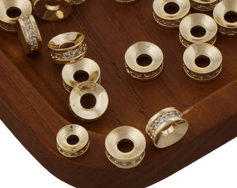 May include: A collection of gold-colored spacer beads with a central hole. Some beads are adorned with a band of small, clear crystals. The beads are arranged on a dark wooden tray, creating a visually appealing display for jewelry making or crafting.