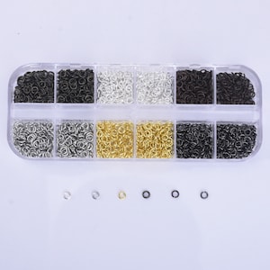 May include: A clear plastic storage box filled with small jump rings in various colors, including silver, gold, black, and bronze. The jump rings are arranged in rows and columns within the box. There are also a few individual jump rings on a white surface below the box.