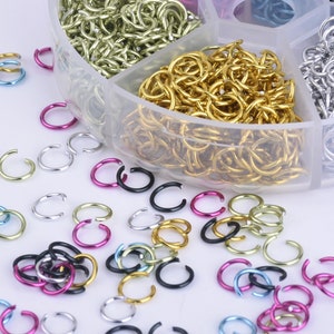 Op de afbeelding: Een doorzichtige plastic container gevuld met verschillende gekleurde metalen springringen. De container heeft zes compartimenten, elk gevuld met een andere kleur springringen. De kleuren omvatten goud, zilver, zwart, roze, blauw en groen. De springringen zijn verspreid rond de container op een wit oppervlak.