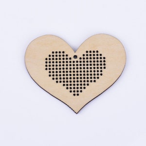 Puede incluir: Un adorno de madera en forma de coraz&oacute;n con un dise&ntilde;o de punto de cruz negro en el centro. El adorno tiene un agujero en la parte superior para colgarlo.