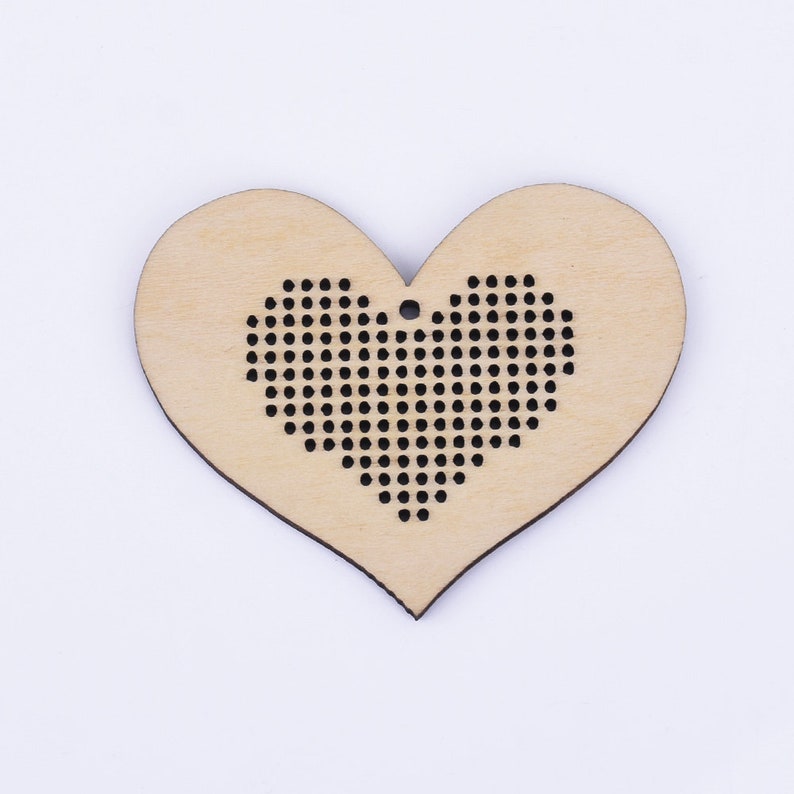 Puede incluir: Un adorno de madera en forma de coraz&oacute;n con un dise&ntilde;o de punto de cruz negro en el centro. El adorno tiene un agujero en la parte superior para colgarlo.
