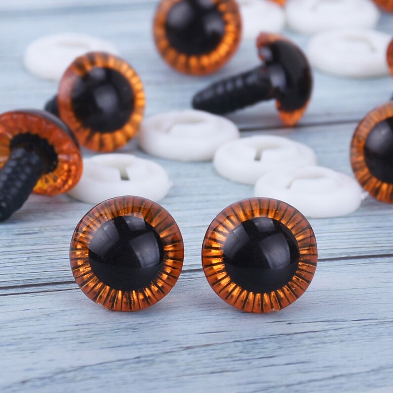 May include: A close-up of two orange and black plastic safety eyes with a shiny finish. The eyes are on a light wood surface.