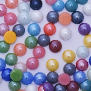 Könnte beinhalten: Eine Sammlung bunter, runder, dekorativer Glassteine in verschiedenen Farben, darunter Rosa, Blau, Grün, Gelb und Orange. Die Steine haben eine glänzende, perlenartige Oberfläche und sind auf einer hellen Oberfläche verstreut. Diese dekorativen Steine eignen sich für Bastelarbeiten und Verzierungen.