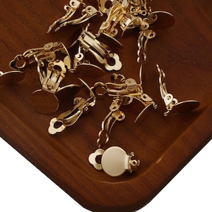 Op de afbeelding: Een close-up van een groep gouden oorbelsluitingen. De sluitingen zijn gemaakt van metaal en hebben een clip-ontwerp. Ze zijn gerangschikt op een houten oppervlak.