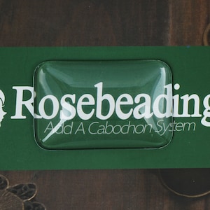 以下が含まれることがあります： 白い文字で「Rosebeading Add A Cabochon System」と印刷された透明なプラスチック製の四角形です。