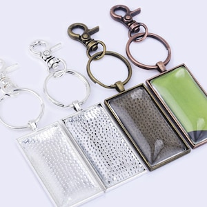 May include: Four keychains with rectangular glass panels. Two keychains have silver metal frames and two have bronze metal frames. The glass panels have a textured pattern. The keychains have a silver, bronze, and copper finish.