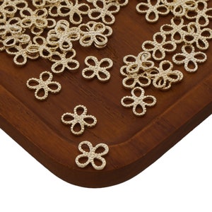 May include: A close-up of a wooden tray filled with gold-coloured metal flower-shaped charms. The charms are small and delicate, with a textured surface.