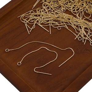 May include: A close-up of a pile of gold-colored earring chains on a wooden surface. Two chains are laid out separately, showing the chain links and the small gold-colored pins attached to the ends.