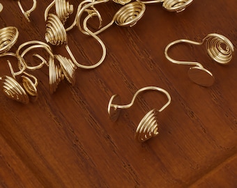 May include: A close-up of a group of gold-toned metal earring findings. The findings are in the shape of a spiral with a small round disc at the end. The findings are scattered on a wooden surface.
