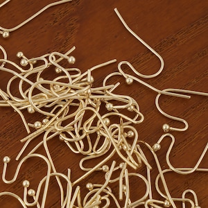 Puede incluir: Un mont&oacute;n de ganchos para pendientes de color dorado con peque&ntilde;as bolas redondas en el extremo. Los ganchos est&aacute;n esparcidos sobre una superficie de madera marr&oacute;n.