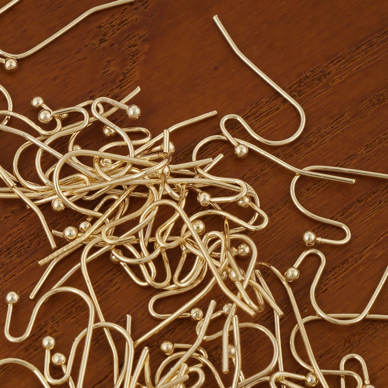 Puede incluir: Un mont&oacute;n de ganchos para pendientes de color dorado con peque&ntilde;as bolas redondas en el extremo. Los ganchos est&aacute;n esparcidos sobre una superficie de madera marr&oacute;n.