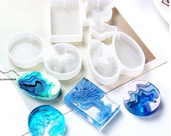 May include: A set of six clear silicone molds for making resin jewelry. The molds are shaped like a square, a crescent moon, a teardrop, a circle, and two abstract shapes. The molds are shown with finished resin pieces in blue and white.