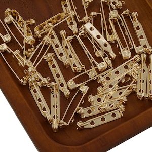 May include: A large quantity of gold-toned metal safety pin backs. The pin backs are arranged in a pile on a wooden surface.