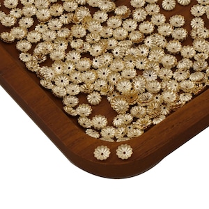 Puede incluir: Una gran cantidad de cuentas de metal de color dorado en forma de flor en una bandeja de madera.