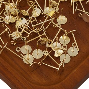 May include: A pile of gold-toned earring posts with loops on the back. The posts are scattered on a wooden surface.