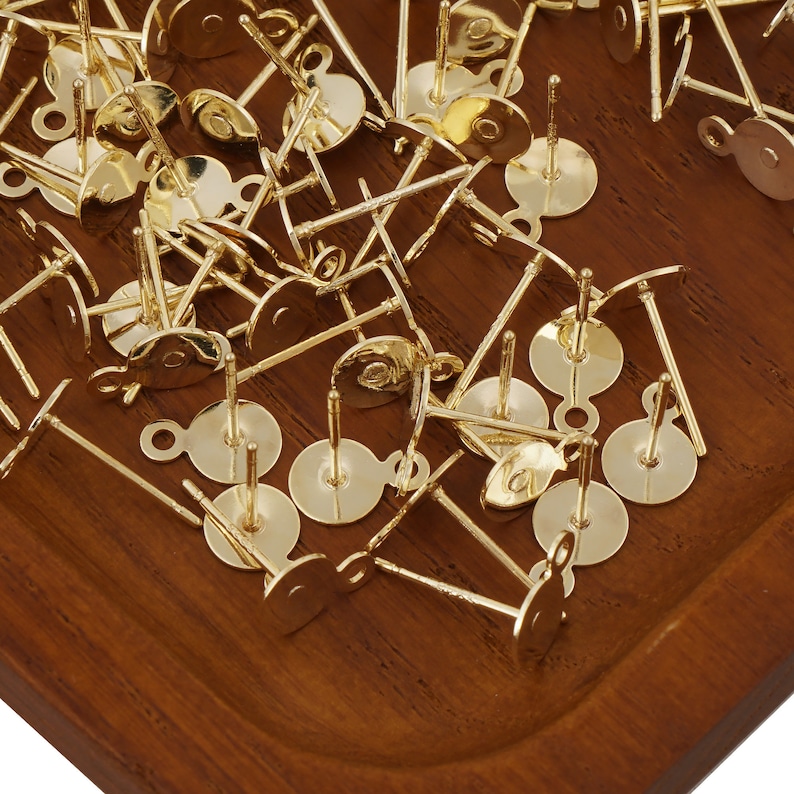 May include: A pile of gold-toned earring posts with loops on the back. The posts are scattered on a wooden surface.