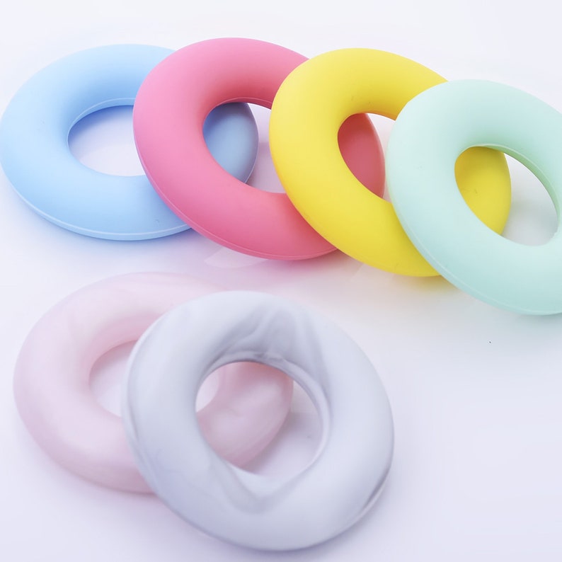 May include: Six colorful silicone rings in various pastel colors, including light blue, pink, yellow, and green. The rings are all in the shape of a donut and are arranged in a semi-circle on a white surface.