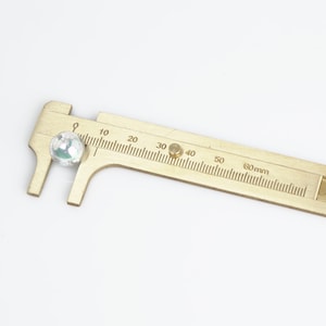 Può includere: Un calibro in ottone che misura una perla di vetro bianca iridescente. Il calibro ha una scala graduata in millimetri fino a 60 mm.