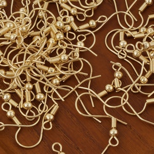 Puede incluir: Primer plano de un mont&oacute;n de ganchos para pendientes de color dorado. Los ganchos son de metal y tienen un peque&ntilde;o bucle en la parte superior y una bola en la parte inferior.