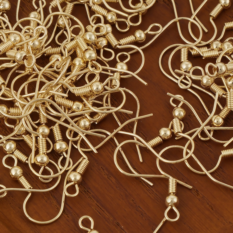Puede incluir: Primer plano de un mont&oacute;n de ganchos para pendientes de color dorado. Los ganchos son de metal y tienen un peque&ntilde;o bucle en la parte superior y una bola en la parte inferior.