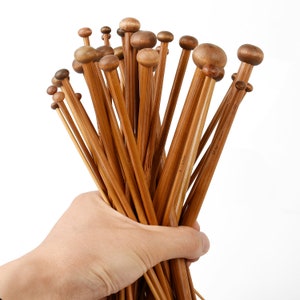 May include: A bunch of wooden knitting needles with natural brown coloured tips. The needles are held in a hand.