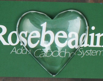 May include: A clear glass heart-shaped cabochon, perfect for jewelry making. The cabochon is sitting on a green background with the text "Rosebeading Add a Cabochon System".